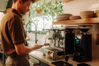 Barista in a brown sweater loads a portafilter on a shiny espresso machine with stacked plates and green plants in the background.