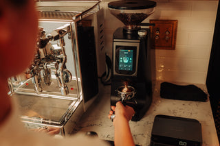 Barista tamping coffee beside a chrome espresso machine and the Eureka Atom 75 grinder with a clear bean hopper on a marble counter.