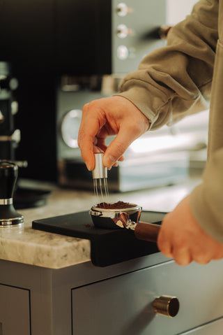 Close up of hand using a four prong distribution tool to fluff coffee grounds in a portafilter