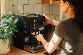 A person uses a black home espresso machine on a wood counter with a green potted plant nearby.