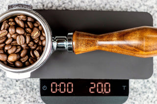 Coffee beans fill a portafilter basket with a wooden handle resting on a digital kitchen scale.
