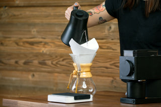 Pouring hot water from a black gooseneck kettle into a paper filter over a pour over dripper above a glass carafe.