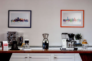 Baratza Virtuoso+ grinder on a kitchen counter with coffee setup and a Moccamaster visible in the background.