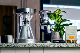Silver automatic drip coffee maker with glass carafe on a speckled countertop beside a coffee grinder and a Verve coffee bag.