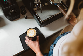 Hands cradle a latte with latte art in front of a polished espresso machine on a white kitchen counter