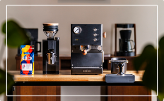 Black espresso machine with gauge beside a tall grinder, a colorful HUCK coffee bag, and a small scale on a wooden counter.