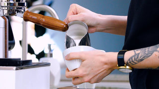 Barista with a tattooed forearm pours steamed milk from a metal pitcher beside an espresso machine.