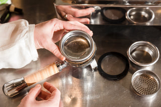 Hands inspect a metal portafilter basket among other tool parts on a stainless counter.