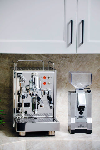 White kitchen cabinets with black bar handles above a stainless espresso machine on a marble countertop.