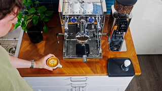 Overhead view of a stainless ECM espresso machine with twin groups and a nearby bean grinder.