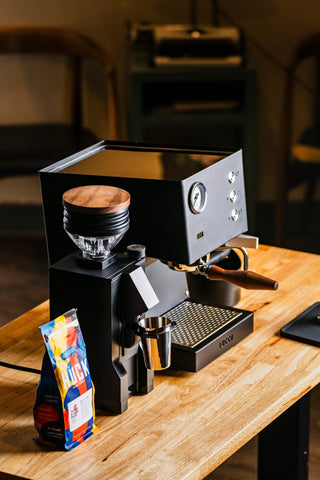 Black espresso setup on a wooden counter with a bag of coffee nearby and a metal portafilter in view