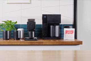 Top-down view of Fellow Aiden Precision Brewer on a wooden counter with a bowl of coffee beans and a digital scale nearby.