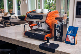 Lucca espresso machine with wooden shell on a marble counter, two white cups on top and a coffee bag nearby.