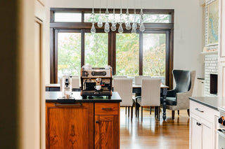 LUCCA espresso machine on a wooden kitchen island in a bright dining area with chairs and large window in the background.