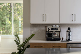Espresso machine on a white kitchen counter beneath upper cabinets with a leafy window view