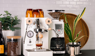 White espresso machine with a clear bean hopper on the left, a glass of espresso on the tray, and a tall grinder on the right