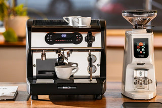LUCCA espresso machine on a wooden counter with a white cup on top and a grinder beside it.