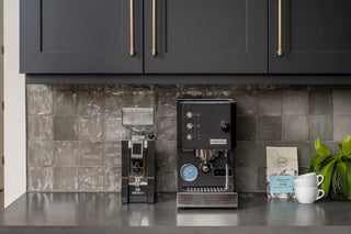 Close up of dark modern cabinetry with brass handles and a black espresso machine beneath against a glossy tiled backsplash.
