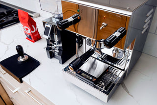 Close up of a polished chrome espresso machine with dual group heads and black handles on a marble counter, a red bag nearby.