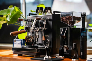 Dual group head espresso machine with wooden portafilter handles and a bean hopper on a counter.