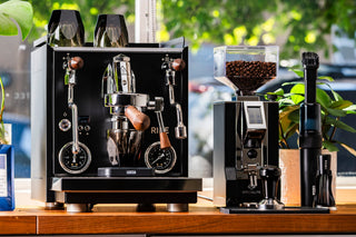 Outdoor coffee setup with a dual head espresso machine on the left and a tall grinder with a bean hopper on the right among greenery