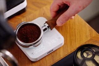 Hand holding a wooden portafilter filled with coffee grounds over a white LUCCA scale on a wooden countertop