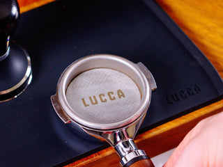 LUCCA espresso portafilter with metal puck screen resting on a black tray