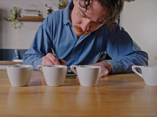 A man in a blue shirt writes at a wooden table with several white coffee cups in the foreground.