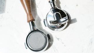 Two portafilters, one with a wooden handle and a chrome one, resting on a light marble surface