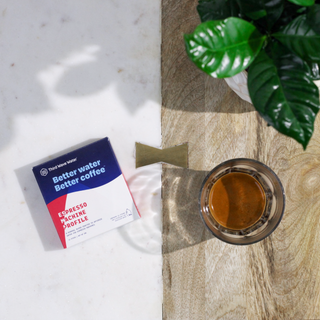 Marble countertop with Third Wave Water packaging on the left and a potted plant on the right near a glass of coffee.