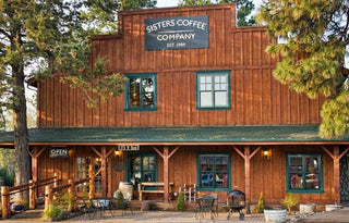 Exterior view of Sisters Coffee Company storefront in a rustic wooden building with green trim and outdoor seating.