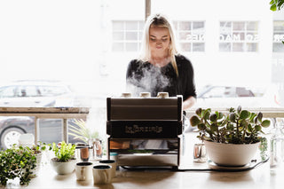 Woman steams milk at a La Marzocco espresso machine on a cafe counter with potted plants nearby.
