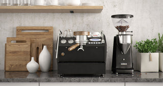 Commercial coffee setup with a large black espresso machine and grinder on a stone counter beside cutting boards and a plant