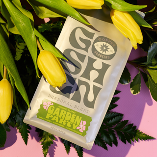 Garden Party Goshen Coffee bag nestled among yellow tulips on a pink surface.
