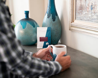A customer holds a cup of coffee beside a Colombian Decaf bag with teal vases in the background.
