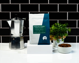 Two Estrada de Terra coffee bags on a counter with a moka pot and a plant against a brick wall.