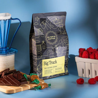 Big Truck coffee bag by Olympia Coffee Roasting Co on a blue background with a blue glass pour over and raspberries.