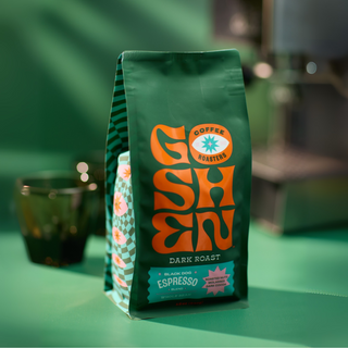Green Black Dog Espresso bag on a green surface with an orange logo and a blurred coffee cup in the background