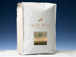 White Kaldi's Coffee bag with a beige patterned side panel and a gold CAFE KALDI label against a deep blue backdrop.
