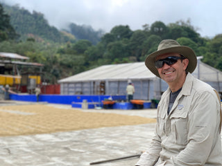 Smiling man in a wide brim hat and sunglasses at a coffee processing site with blue tanks and white canopy structures behind him.