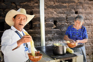 Guatemalan man in a white shirt and straw hat holds a corn cob while a woman husks corn by a pot on a stove in a rustic kitchen.