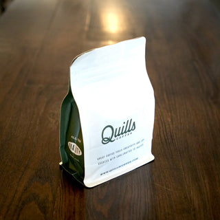 Close-up of a white Quills Coffee bag resting on a dark wooden table.