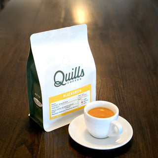 Quills Blacksmith Espresso Blend bag on a wooden table with a white label and a cup of espresso in the foreground
