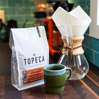 Topeca Breakfast Blend coffee bag on a wooden table beside a glass Chemex with a white paper filter and a green tiled background.