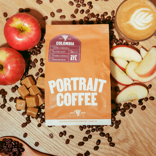 Colombia Sugarcane Decaf bag on a wooden surface surrounded by apples, caramel cubes, and coffee beans with a latte nearby.