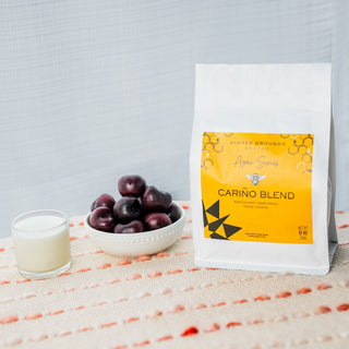Higher Grounds Apex Cariño Blend coffee bag with a bright yellow label placed beside a bowl of grapes and a glass of milk.