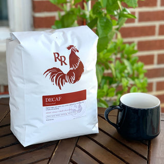 White Red Rooster Coffee bag on a wooden table with green plants in the background and Decaf Seasonal label