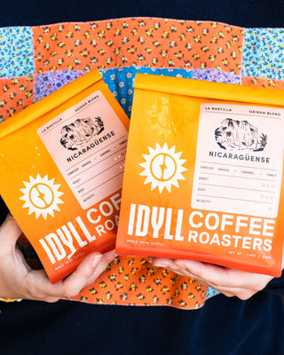 Nicaragüense La Bastilla Geisha Blend coffee bags in bright orange packaging held against a colorful patchwork cloth