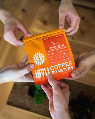 Hands hold a bright orange bag of Togetherness Muungano Cooperative coffee from DR Congo with sun logo.