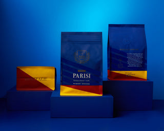 Two Parisi Espresso coffee bags in blue with gold accents on a blue background, accompanied by a yellow and red wrapped package.